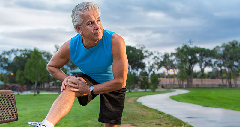 Senior man in a blue tank top stretching his leg outdoors in a park.