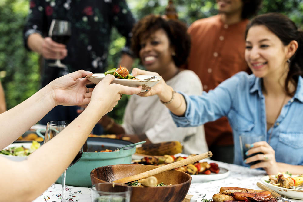Smiling woman in a denim shirt receiving a plate of food at an outdoor gathering with friends