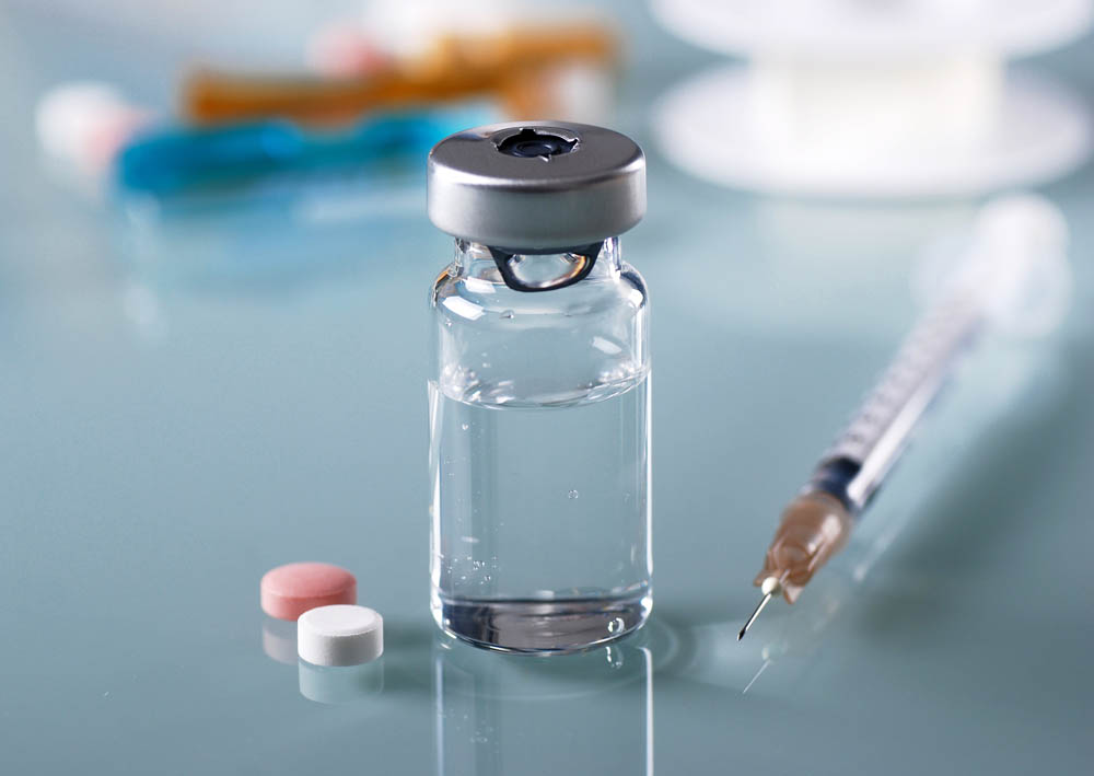 A vial filled with clear liquid, a syringe beside it, and two pills, one pink and one white, on a smooth surface.