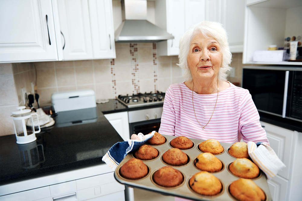 Older woman in a kitchen holding a tray of freshly baked muffins, offering them with a warm expression.