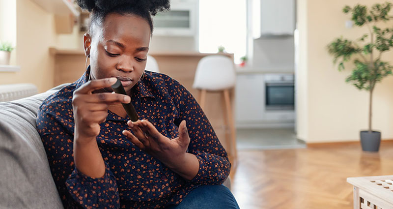 Woman sitting on a couch at home, checking her blood sugar levels with a glucose monitor.