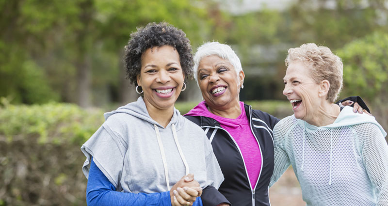 Three older women laughing together outdoors, dressed in athletic wear, enjoying a moment of friendship and wellness.