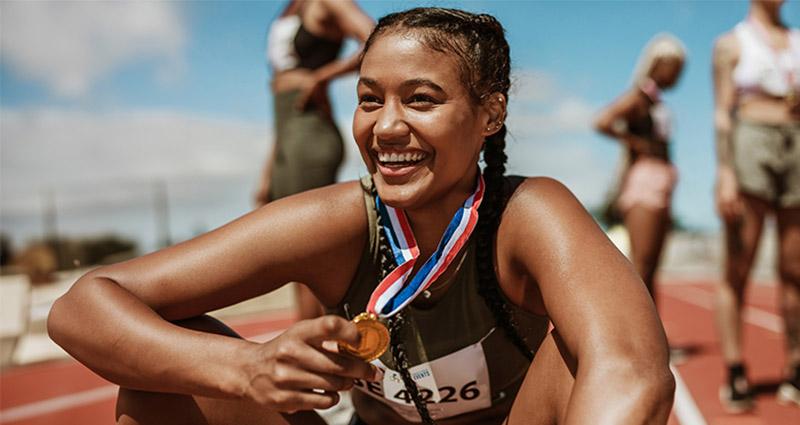 A joyful female athlete sitting on a track, holding a gold medal and smiling after a race, with fellow competitors in the background.