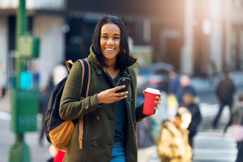 Beautiful latin student woman drinking coffee outdoors on city street and texting on mobile phone. Beautiful young student girl with a bag smiling outdoors.
