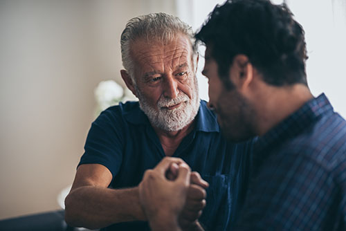 An elderly man with a white beard and a younger man share a heartfelt moment, clasping hands and looking at each other with warmth and understanding.