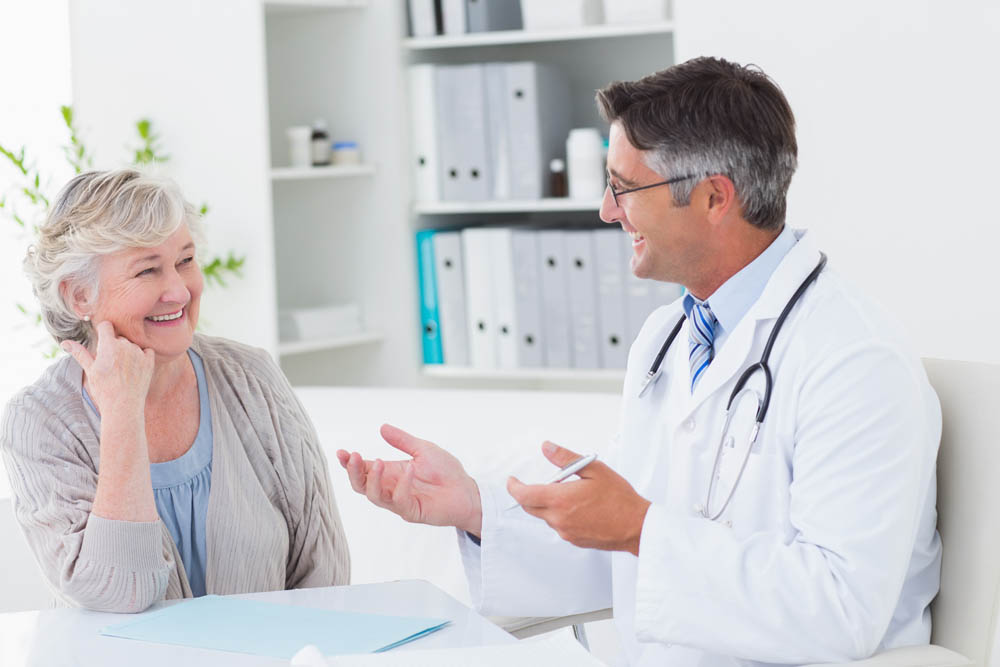 A senior woman smiles while having a conversation with a male doctor in a bright medical office, fostering a positive and reassuring interaction.