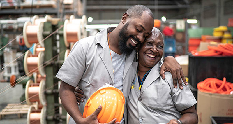   Two workers in a warehouse are smiling and embracing each other, one holding a safety helmet, showcasing a moment of camaraderie and joy in a professional setting.