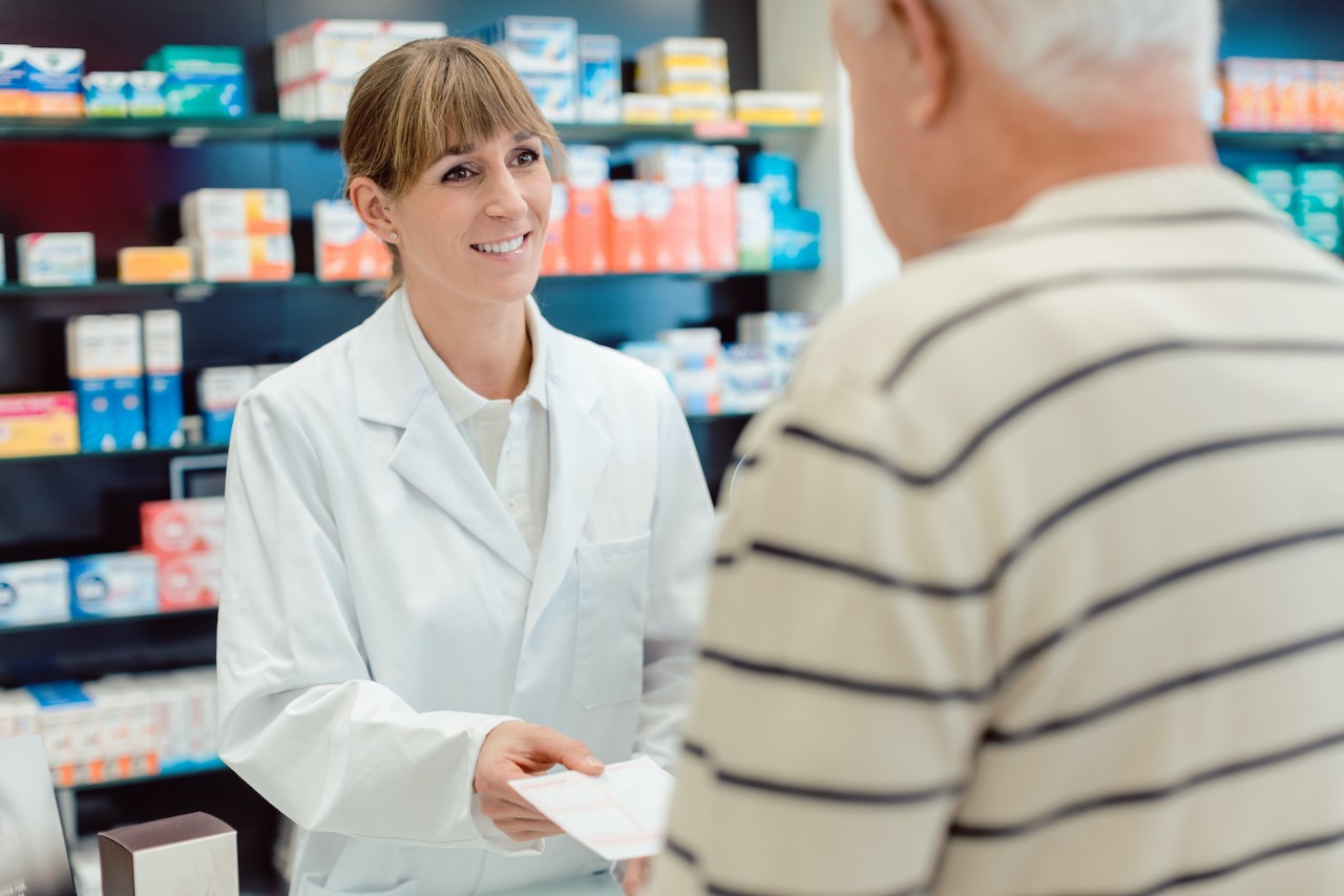 A pharmacist in a white coat handing a prescription to a customer in a pharmacy. Shelves with medical supplies are visible in the background.