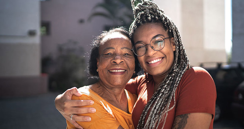 A young woman and an older woman are joyfully embracing each other, both smiling brightly against a sunny background, capturing a warm and loving moment between them.