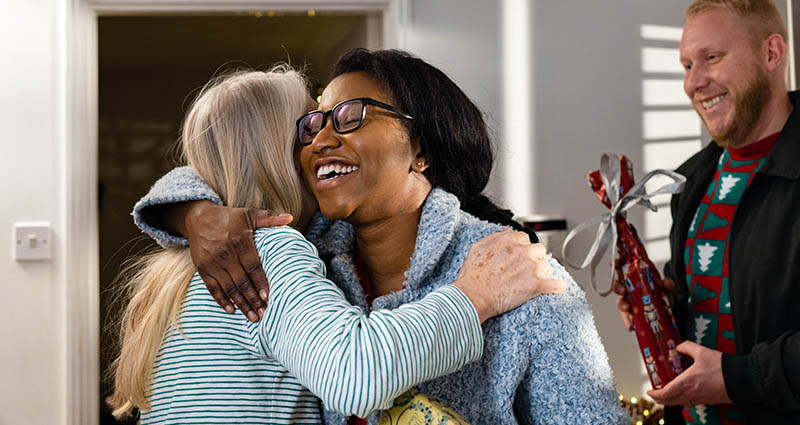 A woman with glasses is happily embracing another woman, while a man in the background smiles and holds a trophy. The scene conveys warmth and celebration in a joyful setting.