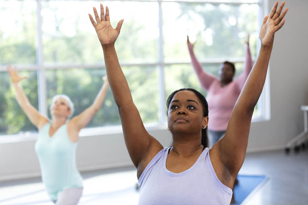 A multi-ethnic group of three women taking a yoga class, in a warrior 1 pose with arms raised. The focus is on the African-American woman in the foreground. She is in her 30s.