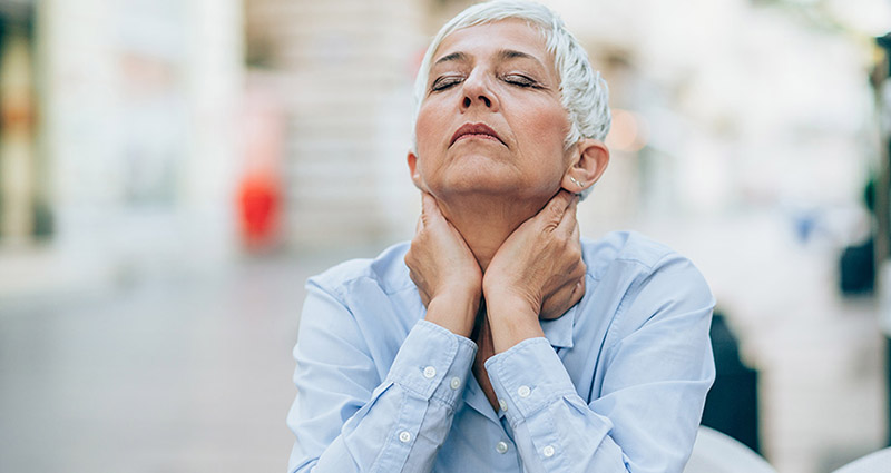 An older woman with short gray hair is sitting outdoors, looking upward with a thoughtful expression, her hands gently resting on her neck, conveying a moment of reflection.