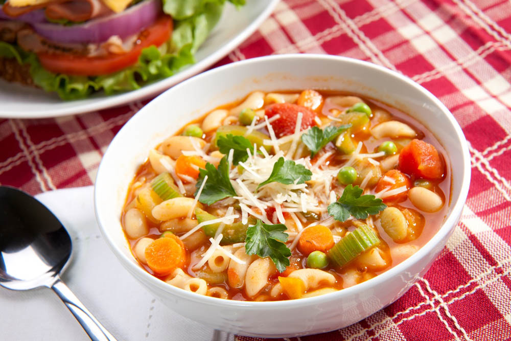 A bowl of vegetable soup filled with beans, corn, and green vegetables, garnished with fresh herbs, placed on a red and white checkered tablecloth, alongside a plate of salad in the background.