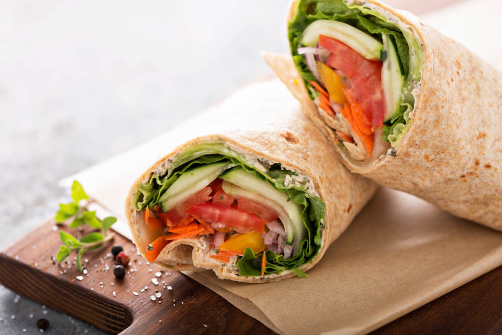 A vegetarian wrap cut in half, filled with lettuce, tomatoes, cucumbers, carrots, and peppers, on a wooden board with seasoning.