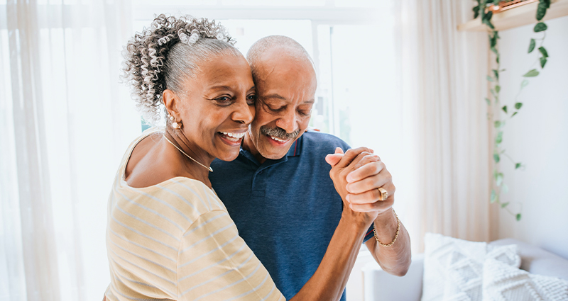 A happy senior couple dances together in a bright living room, holding hands and smiling.