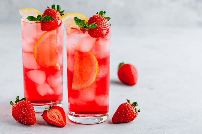 Two tall glasses of strawberry lemonade with ice, garnished with lemon slices, strawberries, and mint, surrounded by whole strawberries on a light surface.