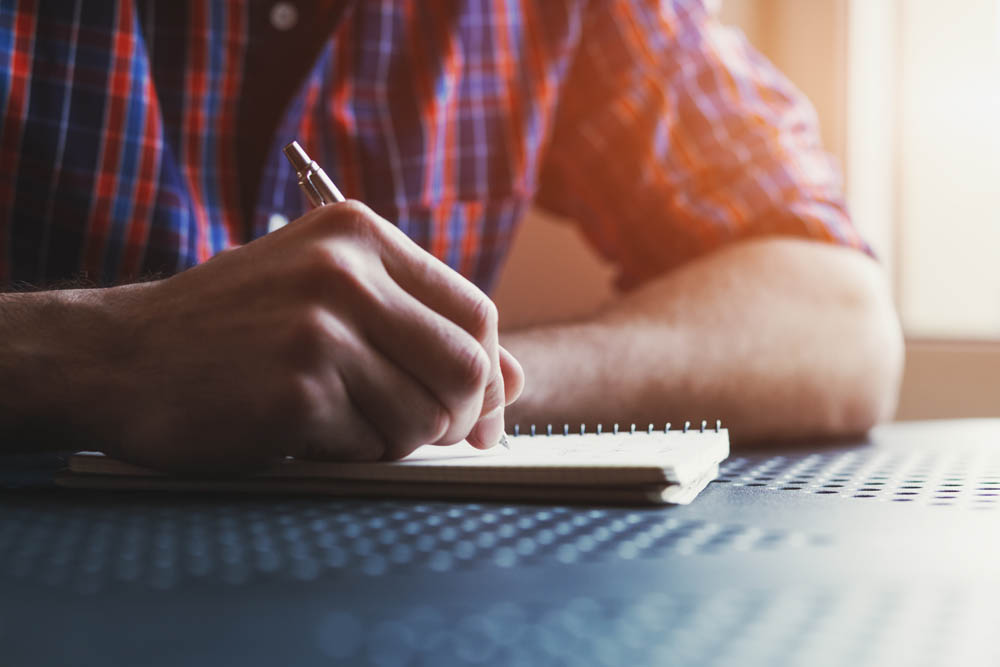 A person wearing a plaid shirt writes in a notebook with a pen, with sunlight streaming in from the side.