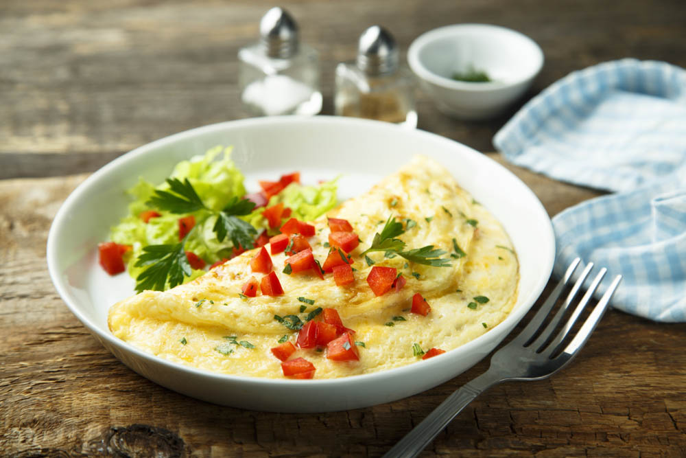 A plate with an omelette topped with diced red peppers and parsley, served with a side of lettuce. Fork and napkin nearby on a wooden table.