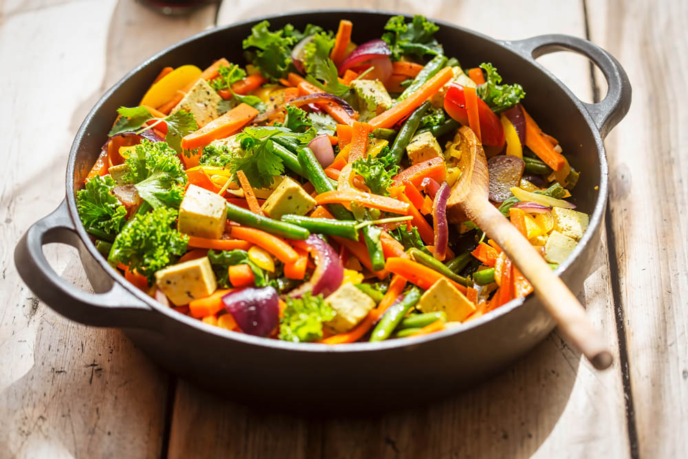 Colorful stir-fry with tofu, broccoli, carrots, peppers, and onions in a black skillet with a wooden spoon, placed on a wooden table.