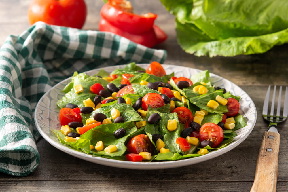 A salad with lettuce, cherry tomatoes, black beans, corn, and red pepper on a plate. A fork and checkered cloth are beside the plate.