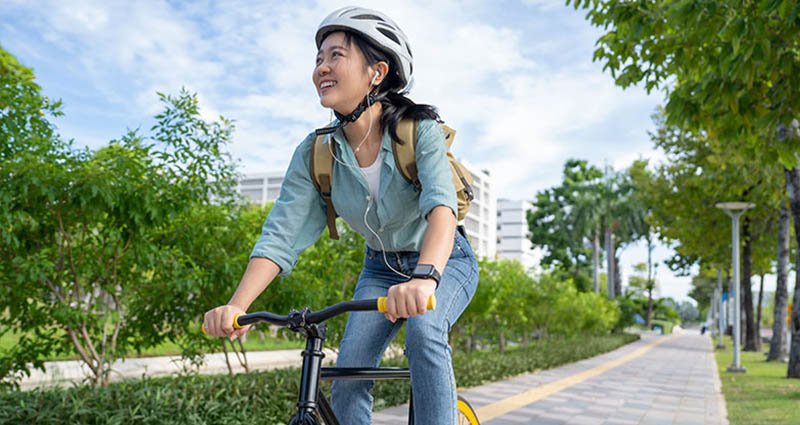 Smiling woman riding a bicycle on a city path with trees, wearing a helmet and backpack.