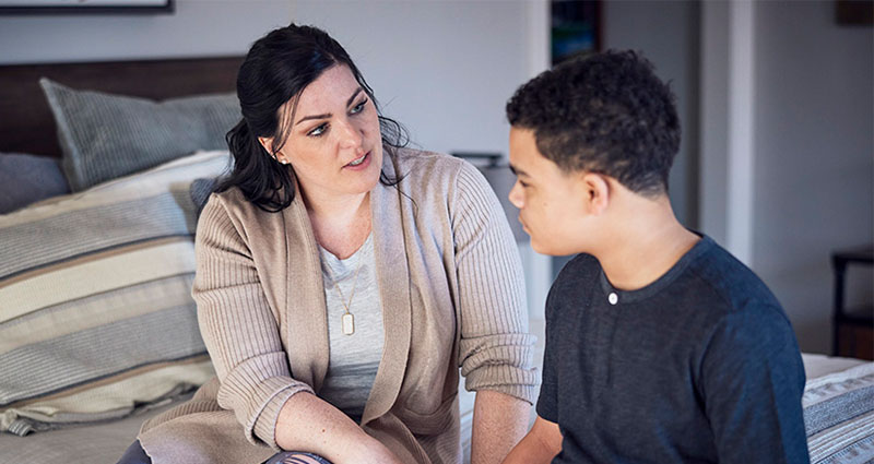 A woman and a young boy are seated on a bed, engaged in a serious conversation, with the woman looking intently at the boy.