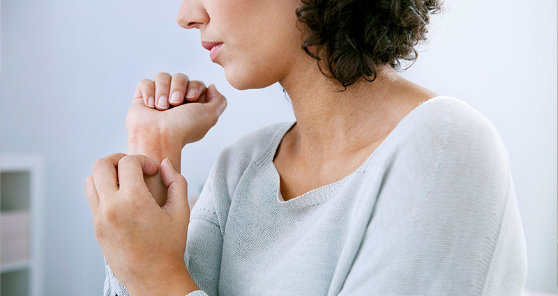  A woman in a light sweater is scratching her wrist, appearing to experience discomfort or irritation.