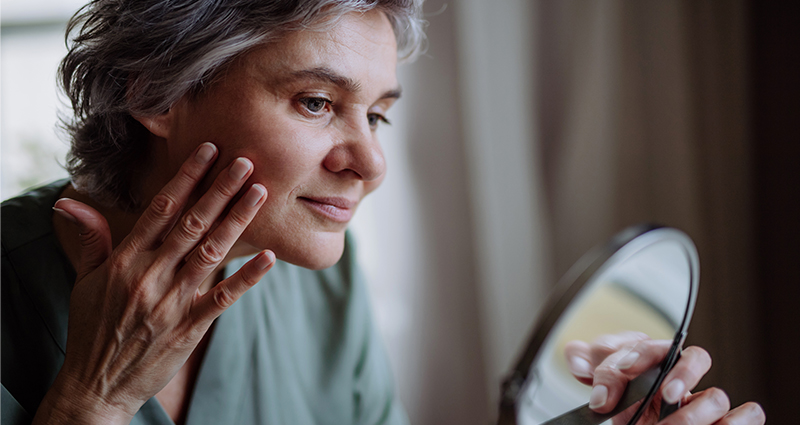 An older woman is examining her face in a mirror, gently touching her skin with a thoughtful expression.