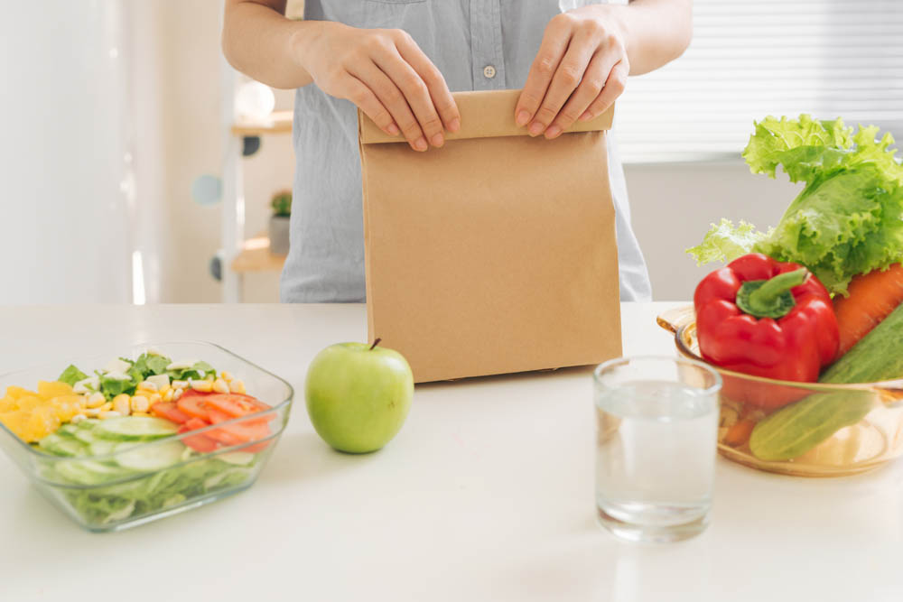 Person packing a brown paper lunch bag, with a salad, green apple, glass of water, and vegetables on the table.