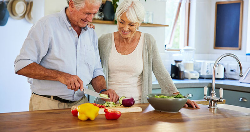 An older couple is preparing food together in a kitchen, with one person chopping vegetables while the other watches, creating a joyful cooking moment.