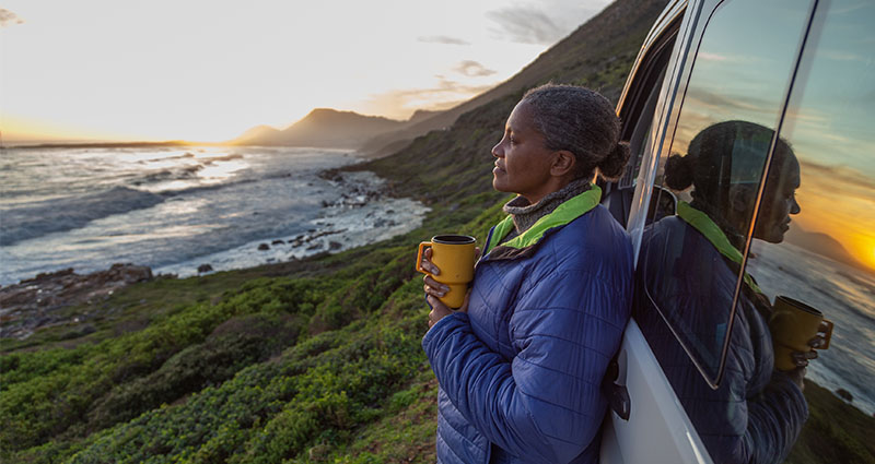 A woman stands by a vehicle, holding a cup and gazing at the ocean and mountains during sunset, reflecting a moment of tranquility and appreciation for nature.