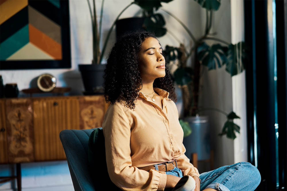 A woman with curly hair is sitting calmly in a chair, eyes closed and appearing to be in a moment of relaxation or meditation, surrounded by indoor plants and a stylish decor.