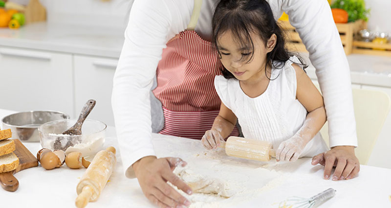 An adult and a child working together in a kitchen, with the child rolling dough on a floured surface, surrounded by baking tools and ingredients.