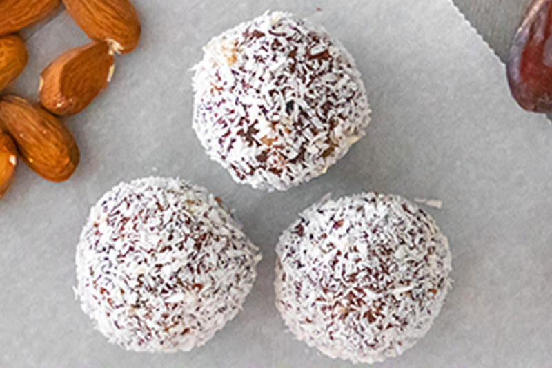 Three coconut-covered brown dessert balls on a white surface, with whole almonds nearby.