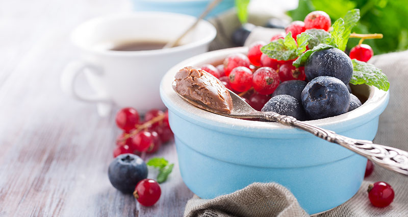 A blue bowl of mixed berries and a spoon with chocolate spread. A cup of coffee is in the background on a wooden table.