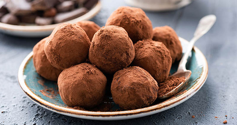 A plate of chocolate truffles dusted with cocoa powder, with a spoon containing cocoa powder beside them, on a textured surface.