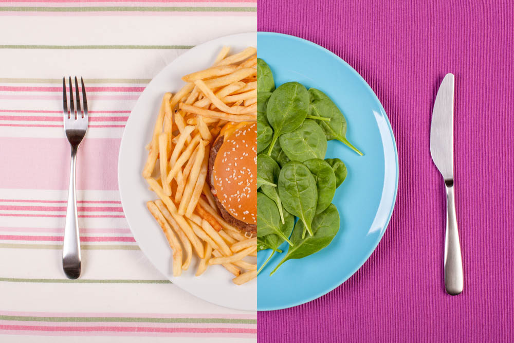 Split image with a burger and fries on the left, and spinach on a blue plate on the right. Fork on the left, knife on the right.