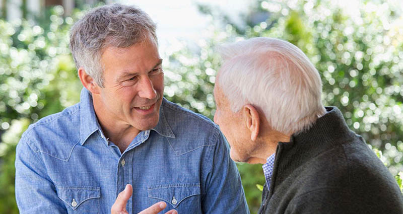 A middle-aged man and an older man are engaged in a lively conversation outdoors, smiling and sharing an animated exchange, with greenery in the background.