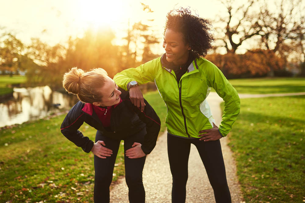 Two women are exercising outdoors in a park during sunset, with one encouraging the other, showcasing camaraderie and support in their fitness journey.