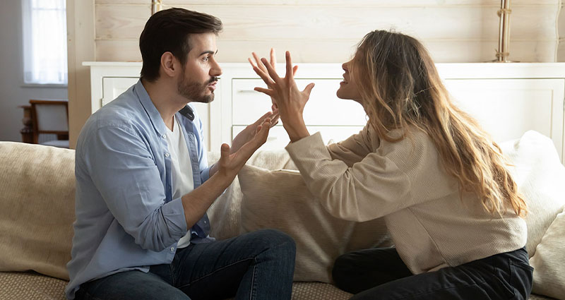 A man and a woman are engaged in a heated discussion or argument while seated on a couch, with expressive hand gestures indicating strong emotions.