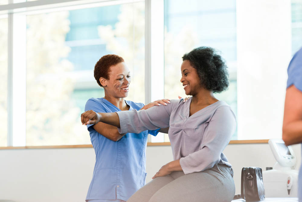As the mid adult female physical therapist massages the mid adult businesswoman's painful shoulder, they both smile.