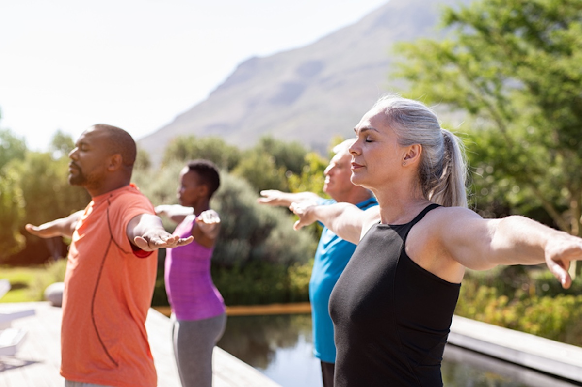 Four people practicing yoga with outstretched arms in an outdoor setting near a pool, with trees and mountains in the background.