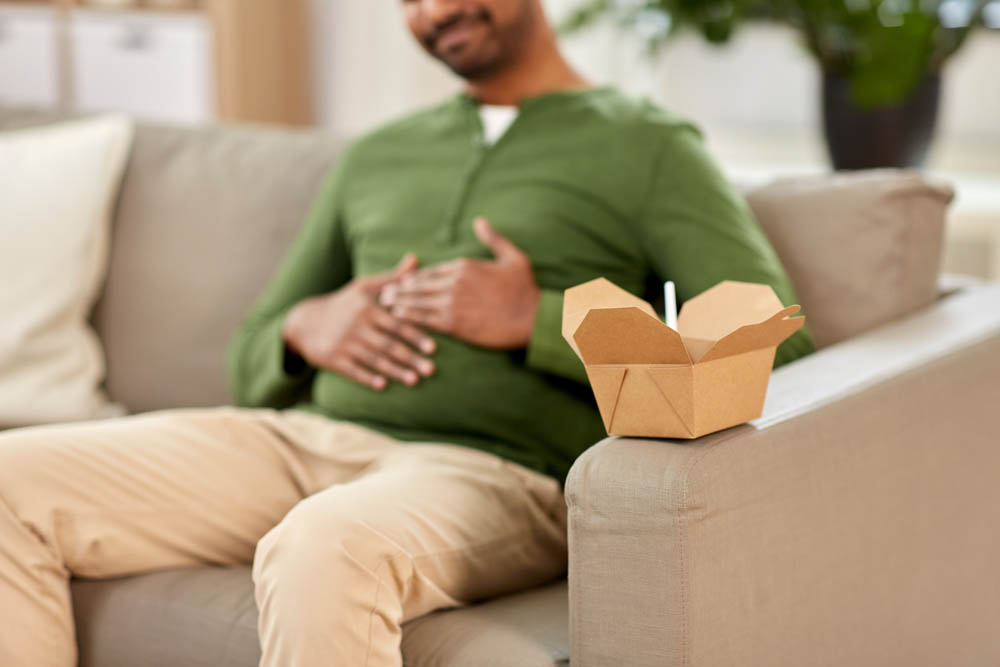 consumption and people concept - cup of takeaway food in front of overeaten man at home