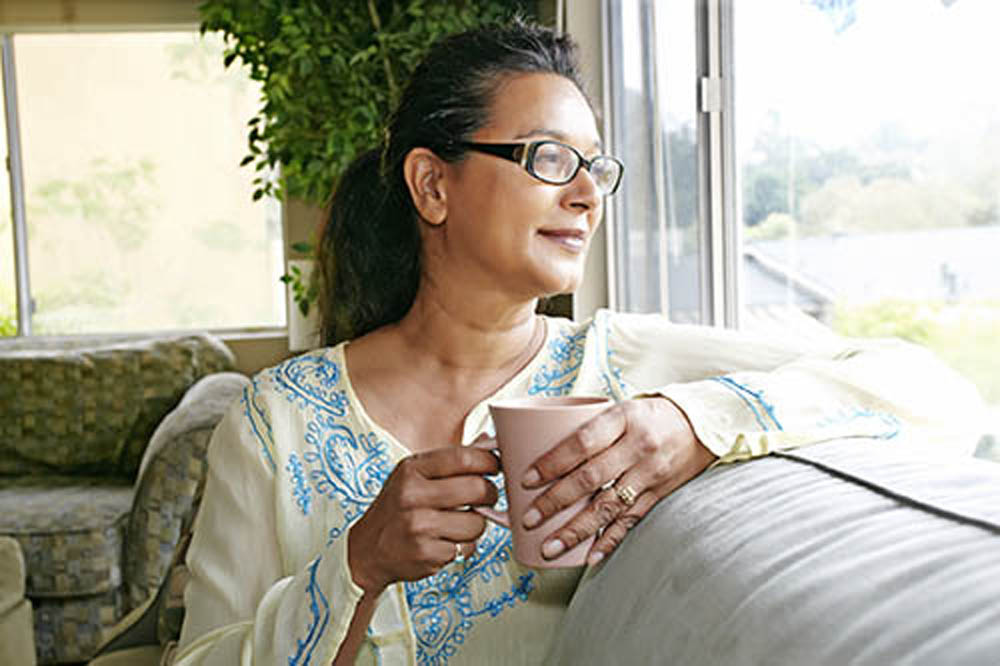 A woman wearing glasses is sitting by a window, holding a cup and gazing thoughtfully outside, surrounded by a cozy indoor setting.