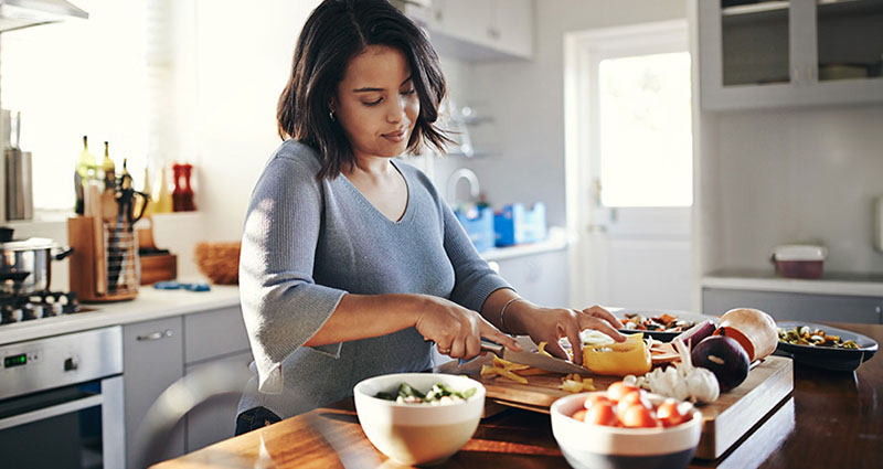 A woman is chopping vegetables in a bright kitchen, focused on her task, with various ingredients and bowls arranged on the countertop around her.