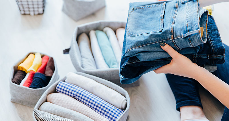A person is organizing clothes, holding a pair of jeans while surrounded by neatly folded items in fabric storage bins.