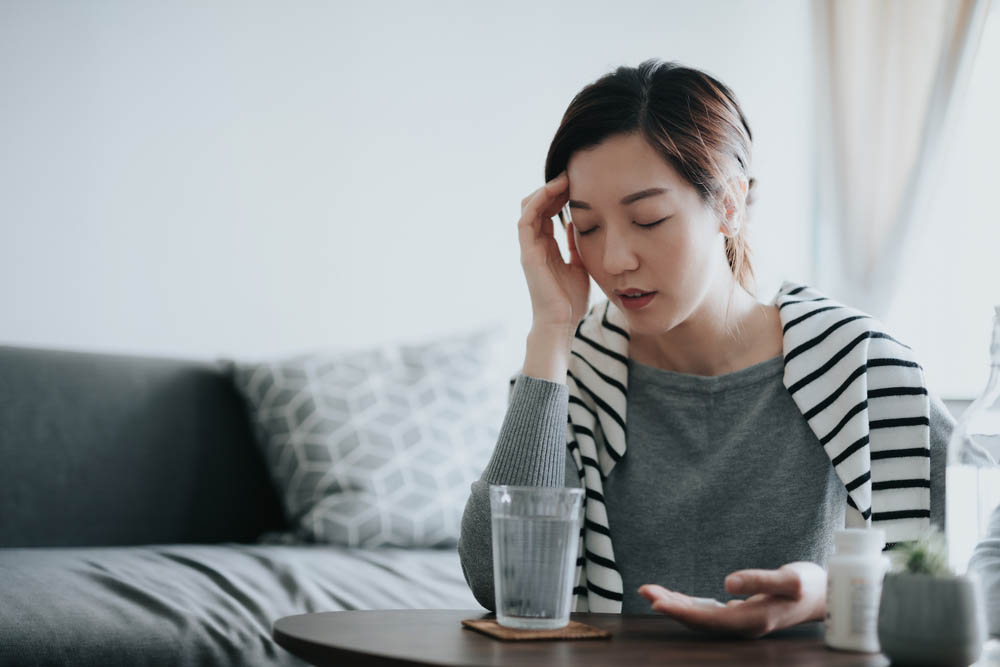 Young Asian woman feeling sick and holding her head in pain, taking medicines in hand, with a glass of water and pill bottle by the side on coffee table. Medicine, healthcare and people concept