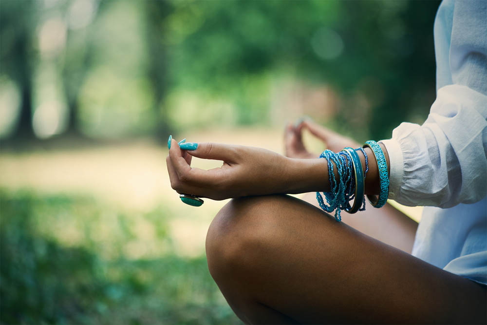  A close-up of a person's hands in a meditative pose, adorned with multiple bracelets, against a blurred natural background.