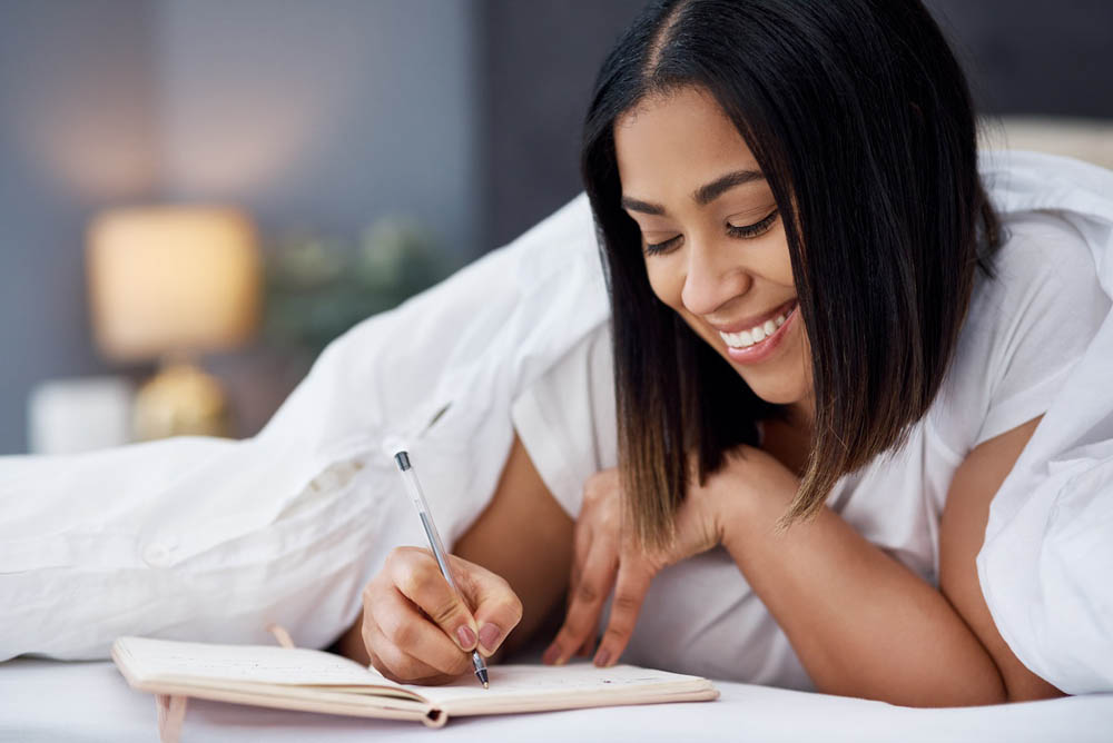 A woman is smiling and writing in a notebook while lying on her stomach in bed, surrounded by soft bedding and warm lighting.