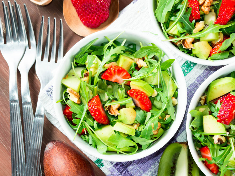 Bowls of salad with arugula, strawberries, kiwi, avocado, and walnuts on a table with forks and a napkin.
